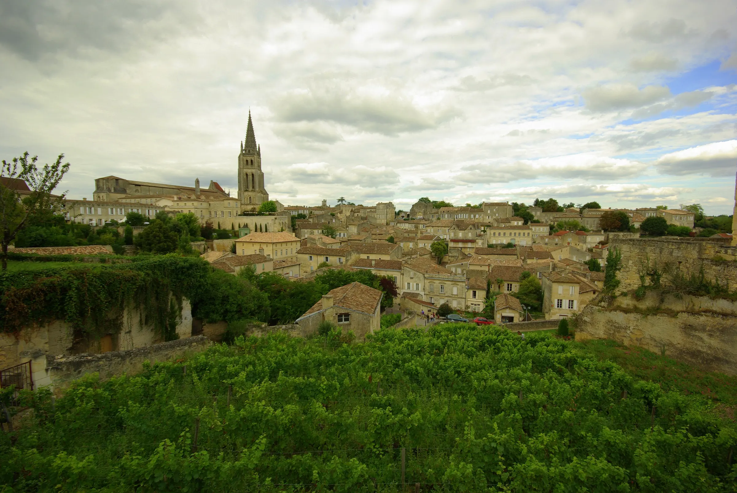 Saint-Émilion rooftops and bell tower rising above a green vineyard under a cloudy sky